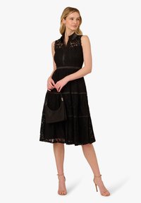Abito nero in pizzo con scollatura a colletto, design senza maniche e gonna a strati. La modella tiene una piccola borsa nera e indossa décolleté color nude.
