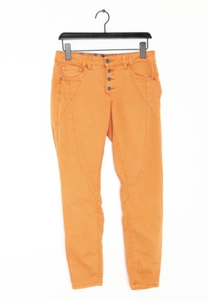 Pantalon orange ajusté avec quatre boutons à l'avant, poches avant et arrière, suspendu à un cintre noir sur un fond blanc.