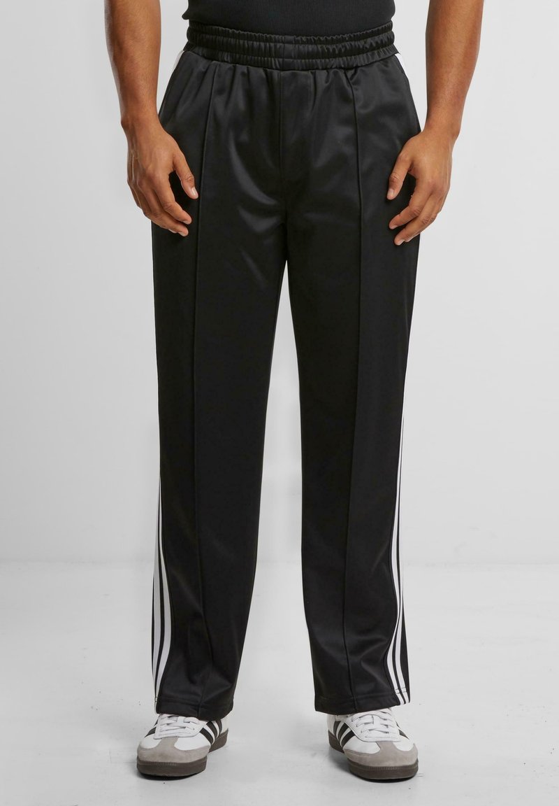 Personne portant un pantalon de survêtement noir avec des bandes blanches sur les côtés et des baskets blanches, debout sur un sol gris clair devant un fond uni.