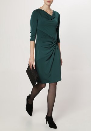 Frau trägt ein dunkelgrünes knielanges Kleid mit geraffter Taille, schwarze transparente Strumpfhosen, schwarze High-Heels und hält eine schwarze Clutch-Tasche.