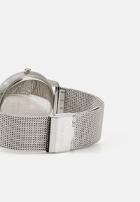 Montre-bracelet en maille argentée avec un boîtier rond en métal, dotée d'un fond en verre, d'un bracelet ajustable et d'une boucle affichant le nom de la marque.