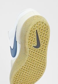 Baskets beige et blanches usées avec un swoosh Nike bleu, montrant une semelle en caoutchouc texturée avec un motif alvéolé et un logo Nike noir.