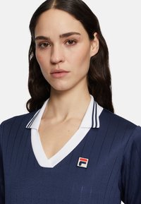 Abito navy con strisce verticali, colletto a V bianco e dettagli a righe. Presenta un piccolo badge con il logo FILA sul lato sinistro. Texture liscia.