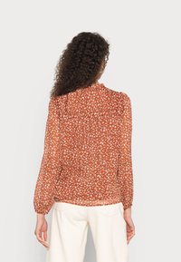 Langärmlige Bluse in verbranntem Orange mit weißem Blumenmuster, durchscheinendem Stoff, gerafften Manschetten und lockerer Passform. Mit einem Knopfverschluss am Rücken.
