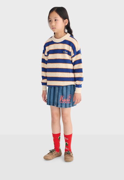 Enfant portant un pull rayé bleu et crème, une jupe en denim bleu avec un texte rouge, des chaussettes rouges à motifs et des chaussures beige, debout devant un fond gris.