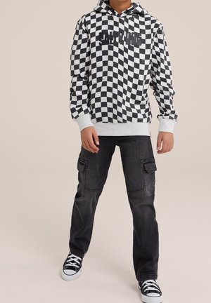 Persona che indossa una felpa con cappuccio a quadri bianco e nero con scritta, pantaloni cargo neri e sneakers nere con lacci bianchi, in piedi contro uno sfondo neutro.