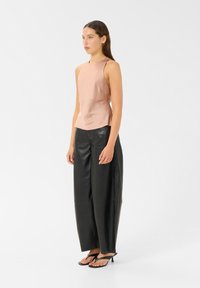 Ljusrosa satin utan ärmar topp, i kombination med löst sittande svarta läderbyxor och svarta flip-flops, stående mot en enfärgad vit bakgrund.