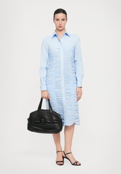 Rochie bleu deschis, cu nasturi, cu detalii din țesătură plisată. Modelul ține în mână o geantă de mână din piele neagră, cu o formă structurată și mânere rulate.