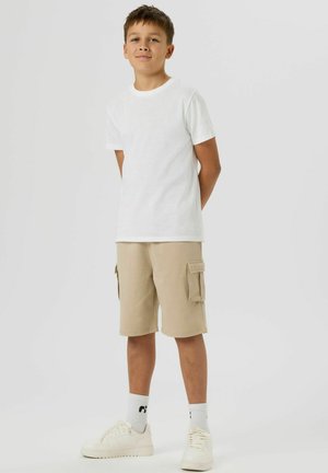 Niño de pie con las manos detrás de la espalda, vestido con camiseta blanca, pantalones cortos cargo beige, calcetines blancos y zapatillas blancas sobre fondo liso.