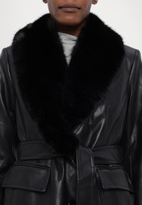 Μαύρ�ο δερμάτινο παλτό με μεγάλο μαύρο γούνινο γιακά, ζωνάκι στη μέση και δύο μπροστινές τσέπες. Λείος υφής με γυαλιστερό φινίρισμα.