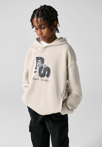Jeune personne aux cheveux tressés portant un sweat à capuche beige Paul Smith avec un motif zèbre et un pantalon noir, les mains dans les poches, regardant vers le bas.