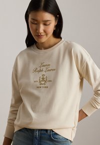 Γυναίκα που φοράει μπεζ φούτερ Lauren Ralph Lauren με λογότυπο και κείμενο, συνδυασμένο με μπλε τζιν, κοιτάζοντας προς τα κάτω με το ένα χέρι στη μέση.