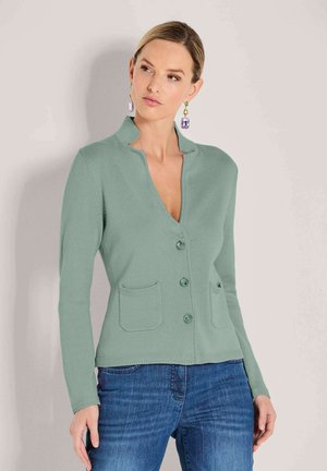 Vrouw in een salie groene gebutton-up cardigan met twee voorzakken en blauwe spijkerbroek, verfraaid met veelkleurige hangende oorbellen.
