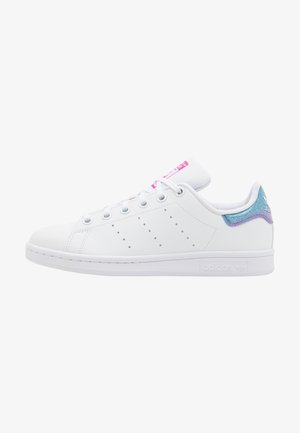 Baskets basses blanches avec bandes latérales perforées, lacets blancs, étiquette rose sur la languette et détail irisé bleu-violet au talon.