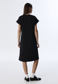 Schwarzes Baumwollkleid mit kurzen Ärmeln, knielang, lockere Passform und glatter Textur. Kombiniert mit schwarzen Schuhen und weißen Socken.