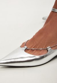 Silberne metallische spitze Ballerinas mit Ausschnitten und einer Kettenakzentuierung um den Knöchel. Glatte Textur und elegantes Design.