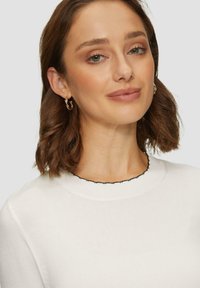 Witte trui met een zwarte golvende halslijn, met een gladde textuur. Model draagt gouden ringoorbellen.