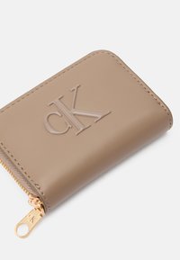 Beżowy portfel z sztucznej skóry z wytłoczonym logo "CK", zamek błyskawiczny oraz złote elementy; gładka tekstura i zaokrąglone krawędzie.