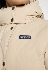 Béžová prešívaná bunda s vysokým golierom, s kovovými cvočkami v striebornej farbe a logom Patagonia v čiernej farbe s modrými akcentmi.