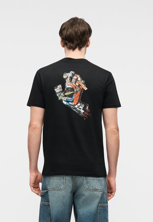 Mies, jolla on musta t-paita, jossa on värikäs grafiikka karjuvasta kädestä, joka pitää tölkkiä ja skeittilautaa selässään, yhdistettynä sinisiin farkkushortseihin.