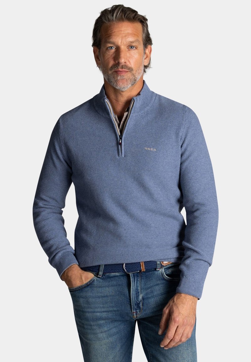 Lichtblauwe, gestructureerde trui met een halve ritssluiting en geribbelde manchetten. Gedragen over een blauwe spijkerbroek, met subtiele branding op de borst.