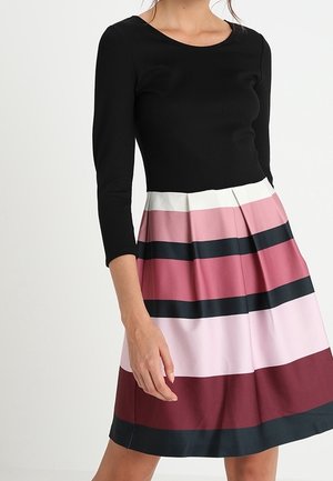 Vrouw draagt een zwarte aansluitende top met driekwart mouwen en een geplooide rok met horizontale strepen in wit, roze, zwart en bordeaux.