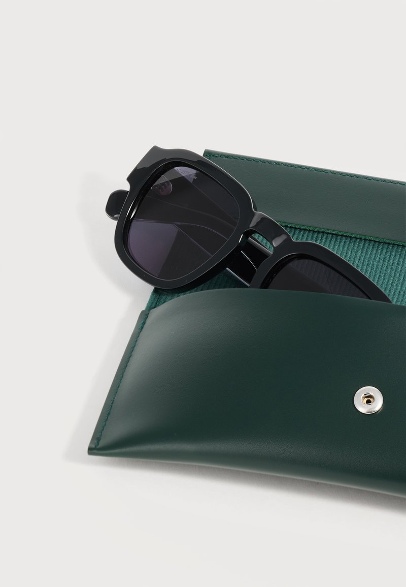 Occhiali da sole con montatura quadrata nera parzialmente all'interno di una custodia in pelle verde scuro aperta con bottone a pressione su sfondo grigio chiaro.