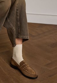 Brown suede loafers with gold studs, paired with a cream ribbed sock, resting on a wooden floor. Trousers have a relaxed fit and muted color.