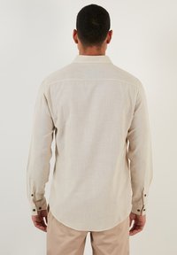 Camisa de manga larga color beige claro, hecha de tela texturizada, con una tapeta de botones, dobladillo redondeado y un solo yugo en la parte trasera.