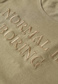 Olivově zelené tričko s vyšitým nápisem "NORMAL IS BORING" v světle béžové barvě. Měkká látka s kulatým výstřihem, minimální textura.