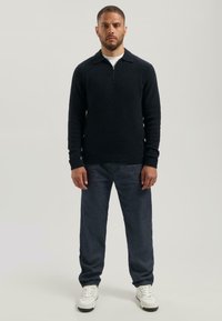 Dunkelblauer Pullover mit Reißverschlusskragen, strukturiertem Stoff und langen Ärmeln, kombiniert mit hellblauen Jeans und weißen Turnschuhen.