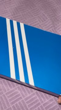 Mėlyna batų dėžutė su blizgiu paviršiumi, viršuje su trimis baltais dantytais dryžiais. Fonas – violetinės lavandos faktūros paviršius.