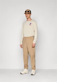 Bežs garās piedurknes krekls ar mazu grafiku, brūna krāsas bikses, baltas kedas un melna ar brūnu cepure un saulesbrilles. Ikdienišķs apģērbs.