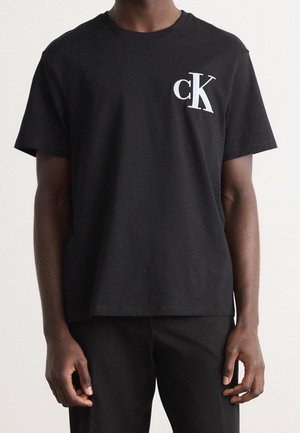 T-shirt en coton noir avec un col rond, des manches courtes, et un logo "ck" blanc contrastant sur le côté gauche de la poitrine. Design simple.