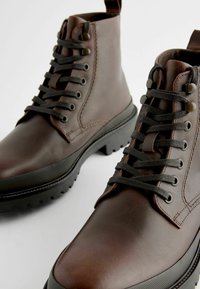 Botas de tobillo de cuero marrón con cordones negros, suela de goma, punta redonda y lengüeta en el talón. Acabado texturizado y detalles de costura reforzada.