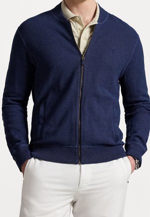 Homme portant un pull zippé bleu marine par-dessus un polo beige avec un pantalon blanc, les mains dans les poches et une montre-bracelet argentée au poignet gauche.