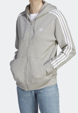 Osoba ubrana w jasnoszarą bluzę Adidas z kapturem na zamek błyskawiczny, z białymi paskami na rękawach, ręce w kieszeniach, połączona z niebieskimi dżinsami.