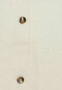 Lichtbeige stoffen shirt met een diagonale weeftextuur, twee grote metalen knopen en een borstzak aan de linkerzijde.