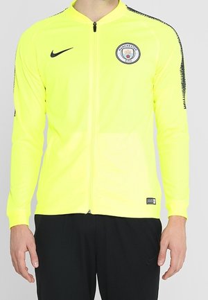 Neonowo żółta kurtka treningowa Manchester City z czarnym logo Nike, zamkiem z przodu i wzorzystymi czarnymi paskami na rękawach.