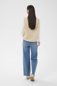 Femme aux longs cheveux lisses et foncés portant un gilet beige sans manches, une chemise blanche à manches longues, un jean bleu et des baskets beiges, debout de dos.