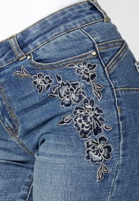 Jean en denim bleu foncé avec un motif de broderie florale sur le côté droit. Comprend un bouton et des rivets en métal, ainsi qu'une texture usée.