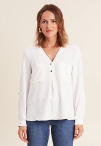 Chemise blanche à manches longues avec décolleté en V, deux poches avant et accents de boutons sur les manches. Fabriquée en tissu lisse et léger.