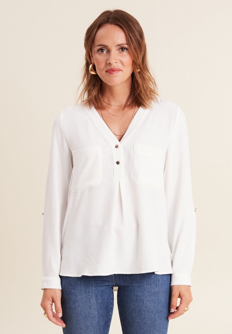 Chemise blanche à manches longues avec décolleté en V, deux poches avant et accents de boutons sur les manches. Fabriquée en tissu lisse et léger.