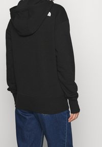 Svart huvtröja med långa ärmar, ribbade manschetter och nederkant. Har en liten vit logotyp på ryggen nära axeln.