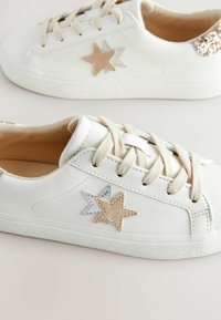 Baskets en cuir blanc avec des appliqués en étoile beige et métalliques, semelle texturée et lacets beige clair. Doublure intérieure en tissu doux.