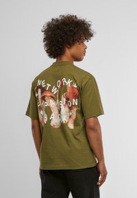 Camiseta verde oliva de corte relajado, con un gran gráfico de champiñones y el texto "PACK DE EXPANSIÓN DE RED" en la parte posterior.