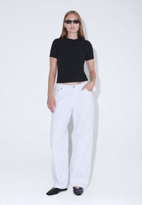 Top negro ajustado de manga corta combinado con jeans de pierna ancha blancos de cintura alta. Modelo llevando gafas de sol oscuras y zapatos negros, fondo neutro.