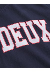 Navy zils audums, uz kura redzama vārds "DEUX" baltās burtos ar sarkanām kontūrām. Tekstūra izskatās gluda ar ikdienišķu dizainu.