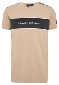 Béžové tričko s černým vodorovným pruhem a bílým nápisem "Kings Will Dream", krátké rukávy a černobílé akcenty na ramenou.