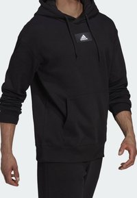 Uomo che indossa un hoodie nero Adidas con tasca frontale, cordini e logo ricamato sul petto, abbinato a pantaloni neri su sfondo neutro.